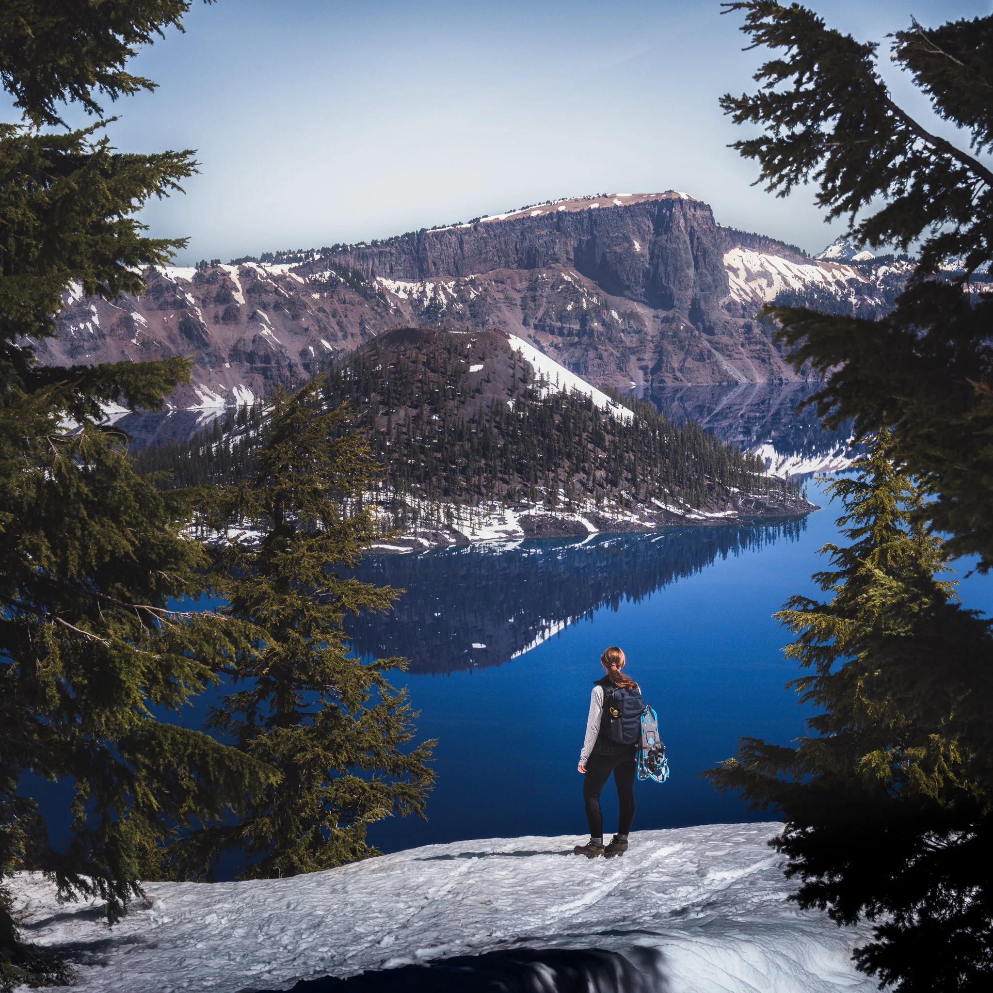 A hiker stands on snow-covered ground near a lake, surrounded by pine trees, with mountains and a clear sky in the background.