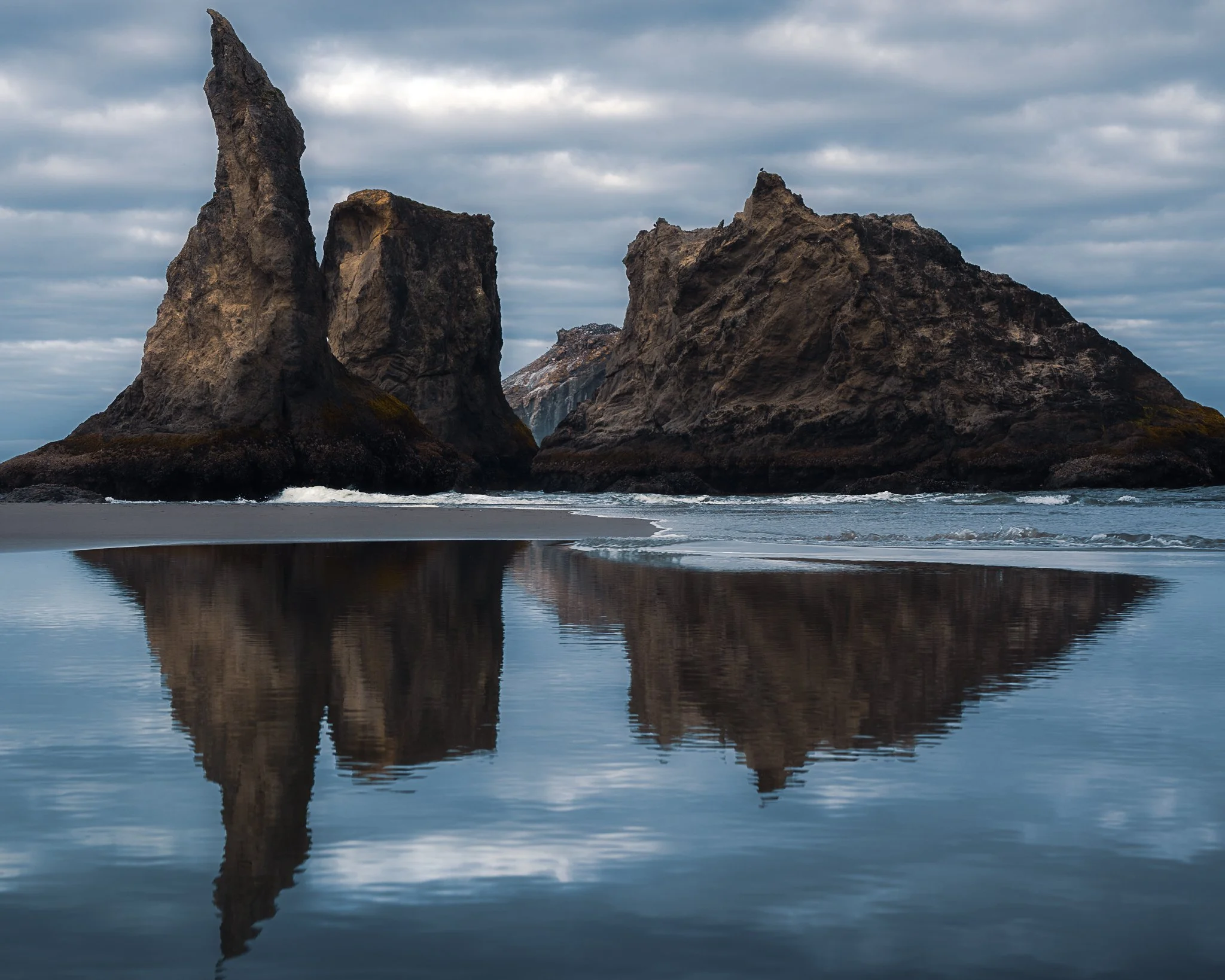 Two large sea stacks on the shoreline with their reflections visible in the wet sand, under a cloudy sky.
