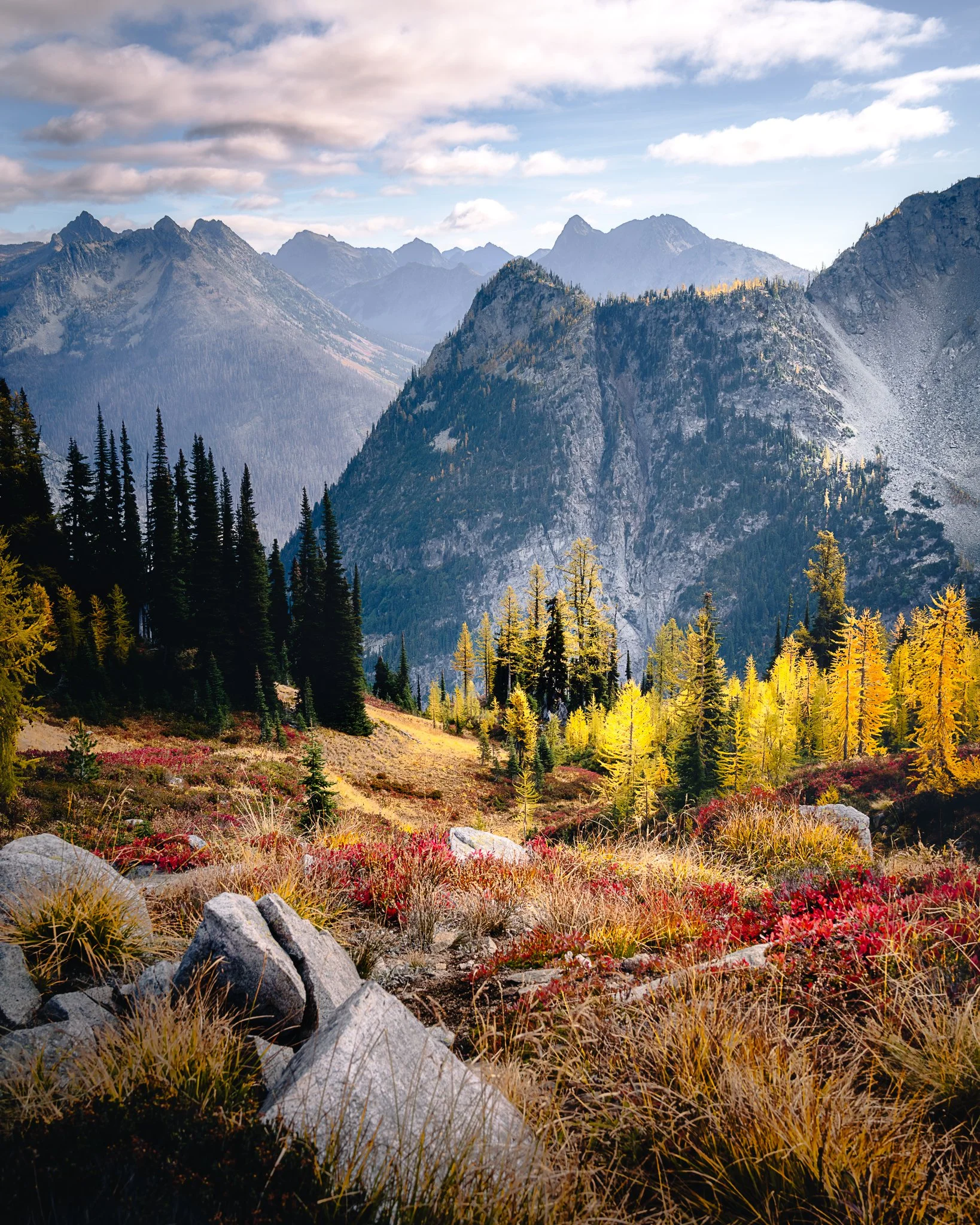 A scenic mountain landscape with forested slopes, rocky peaks, and colorful fall foliage in the foreground under a partly cloudy sky.