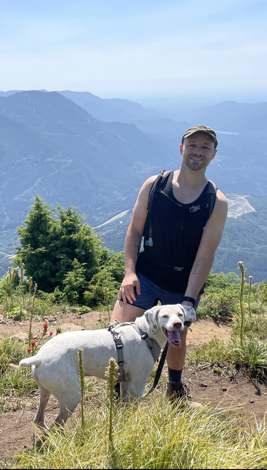 A man with a beard, wearing a black tank top, shorts, and a hat, smiling and posing with a white dog on a mountain trail with a scenic view of mountains and valleys in the background.
