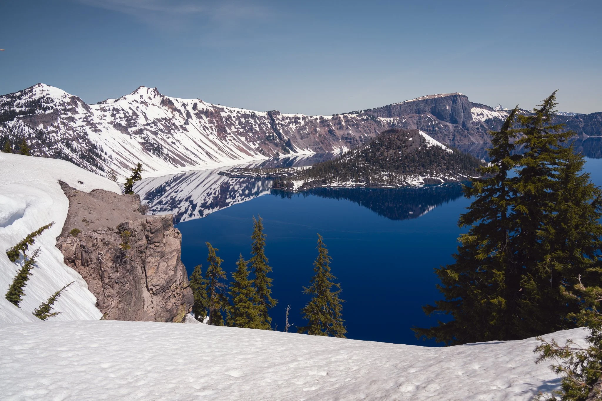 Snow-covered landscape with a large, icy lake reflecting snow-capped mountains and pine trees, under a clear blue sky.