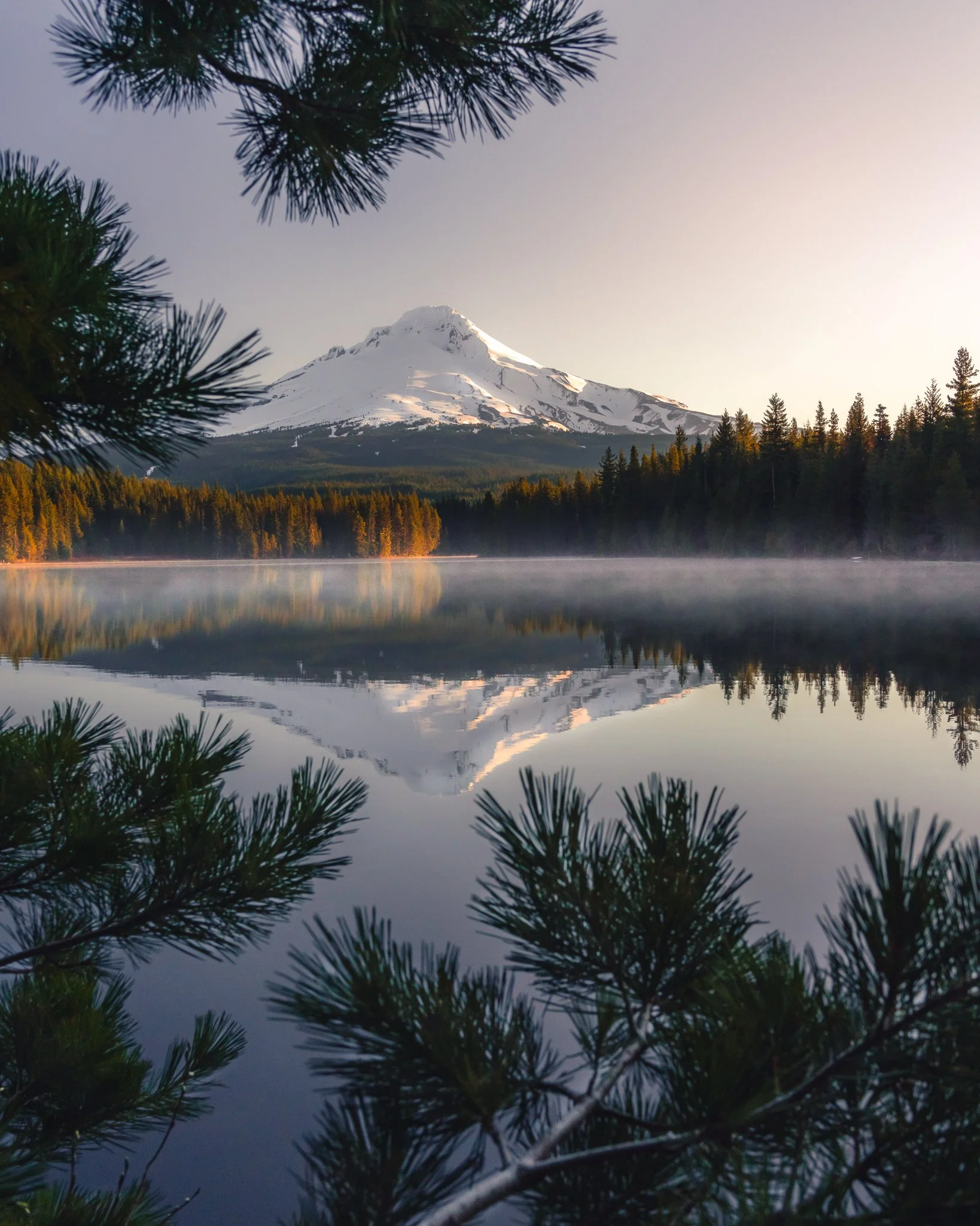 Snow-capped mountain reflected in a calm lake, with trees in the foreground and a clear sky.
