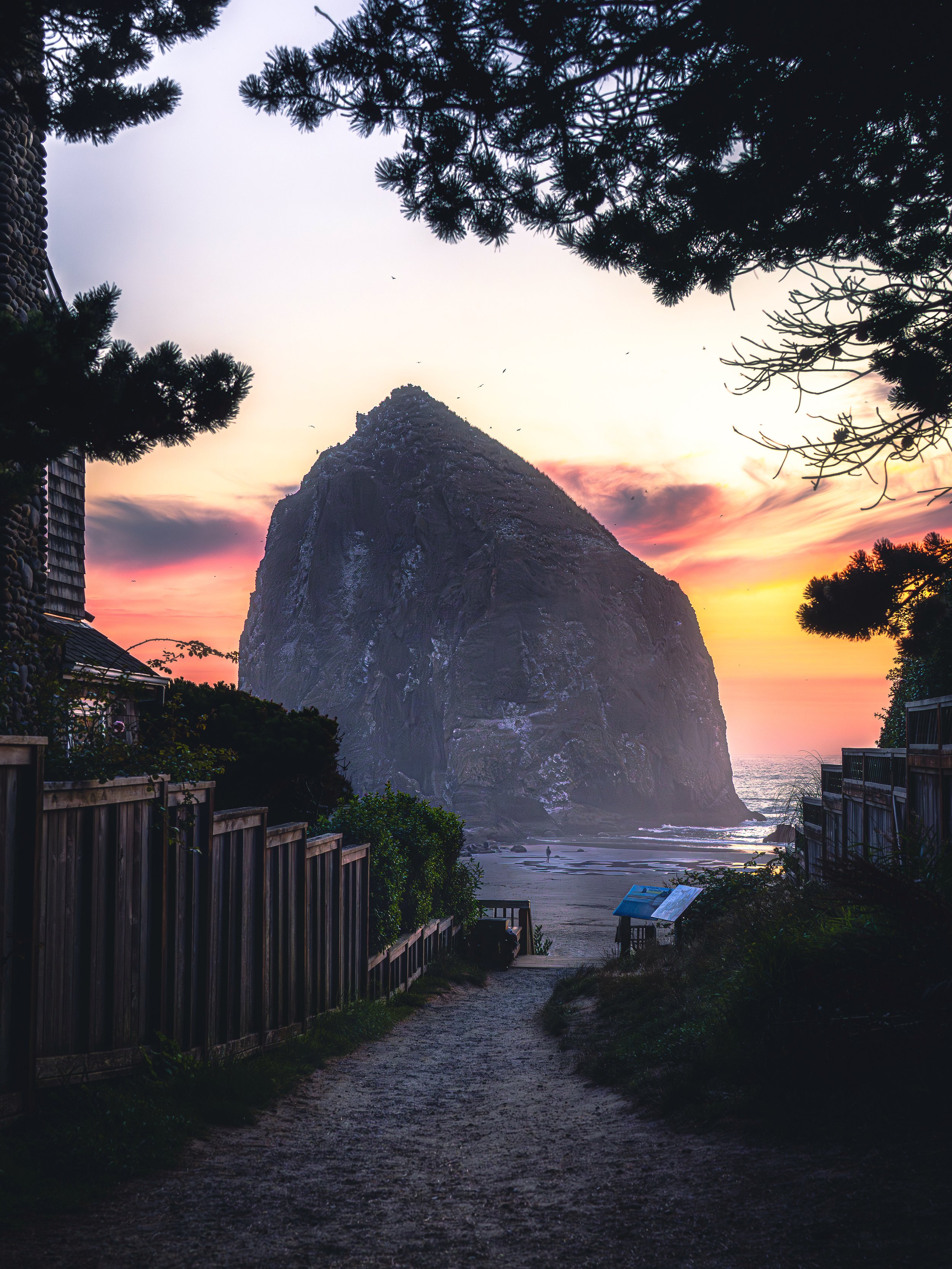Sunset view of a large rock formation on the beach, framed by trees and fences.