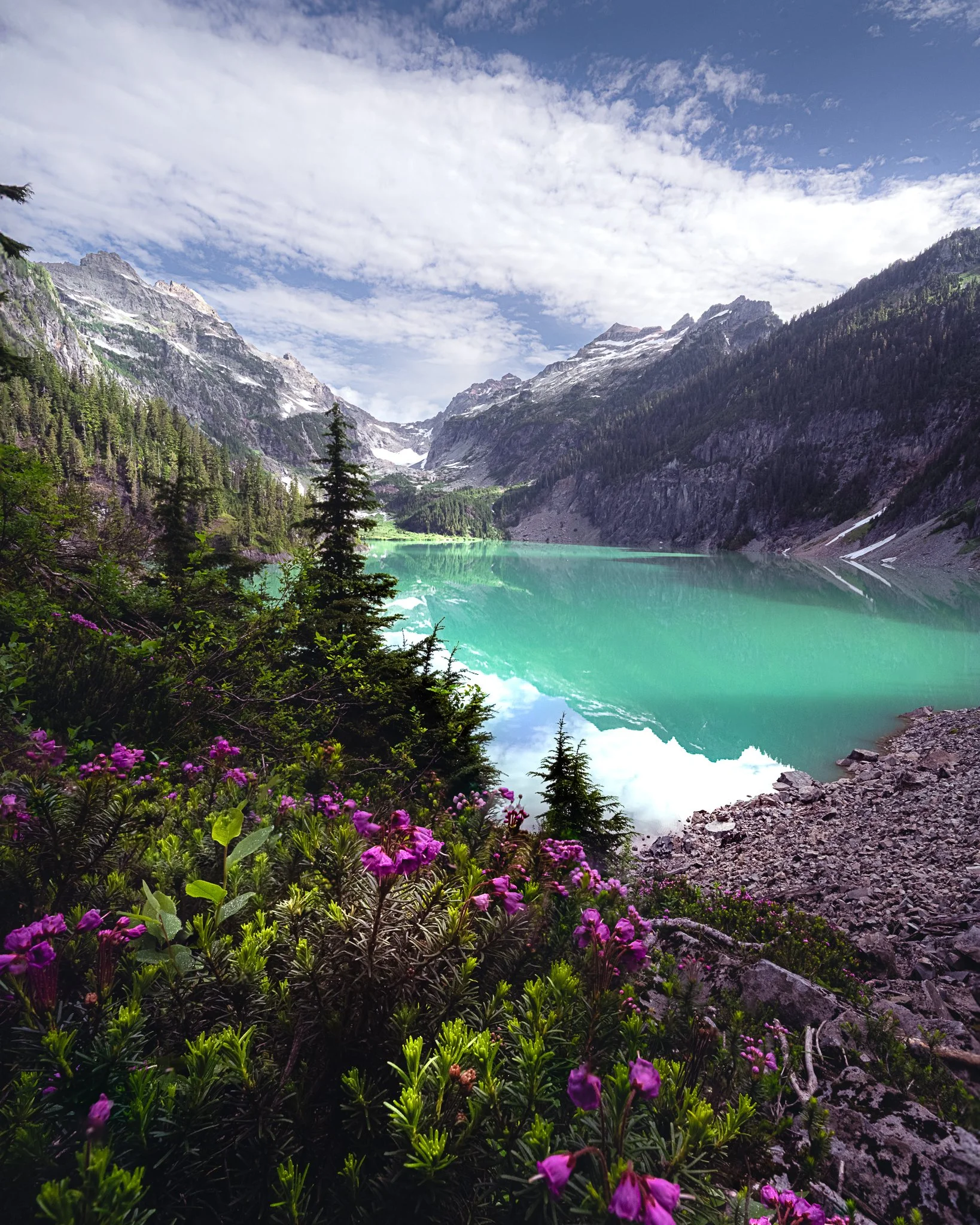 Scenic mountain landscape with a turquoise lake surrounded by evergreen trees, rugged rocky shores, and snow-capped peaks under a cloudy sky.
