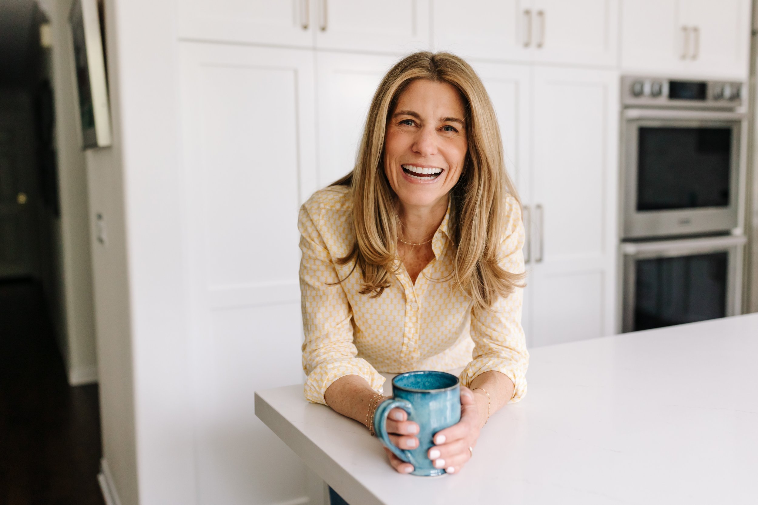 ene Schaffer smiling in a bright, modern space with a blue ceramic mug — reflecting her warm, approachable presence as a coach and speaker focused on clarity, connection, and personal growth.