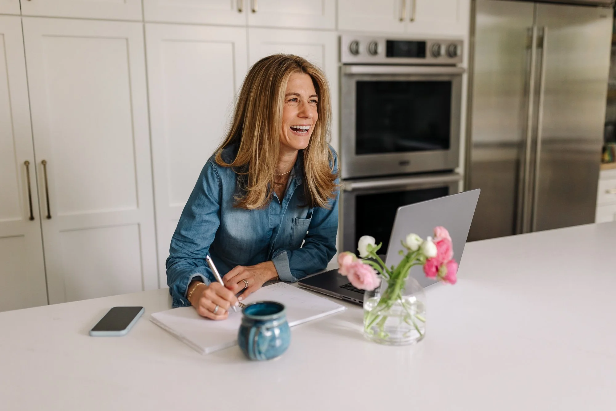 Ilene Schaffer writing at her kitchen table with flowers and tea — reflecting her grounded, home-based coaching style rooted in clarity, warmth, and intentional living.