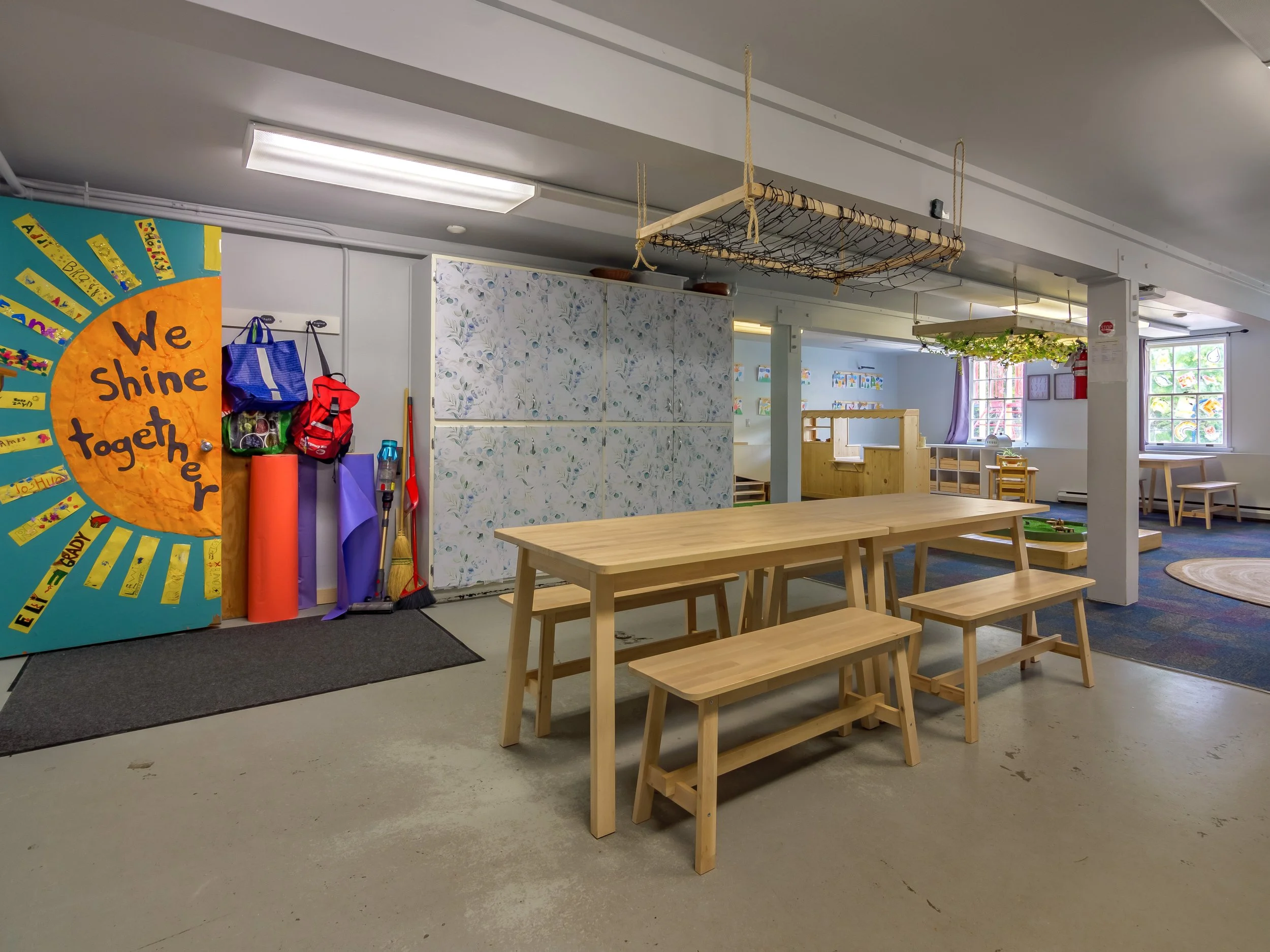 Bright classroom with a wooden table and benches, colourful window decorations, and a wall display with children's artwork.