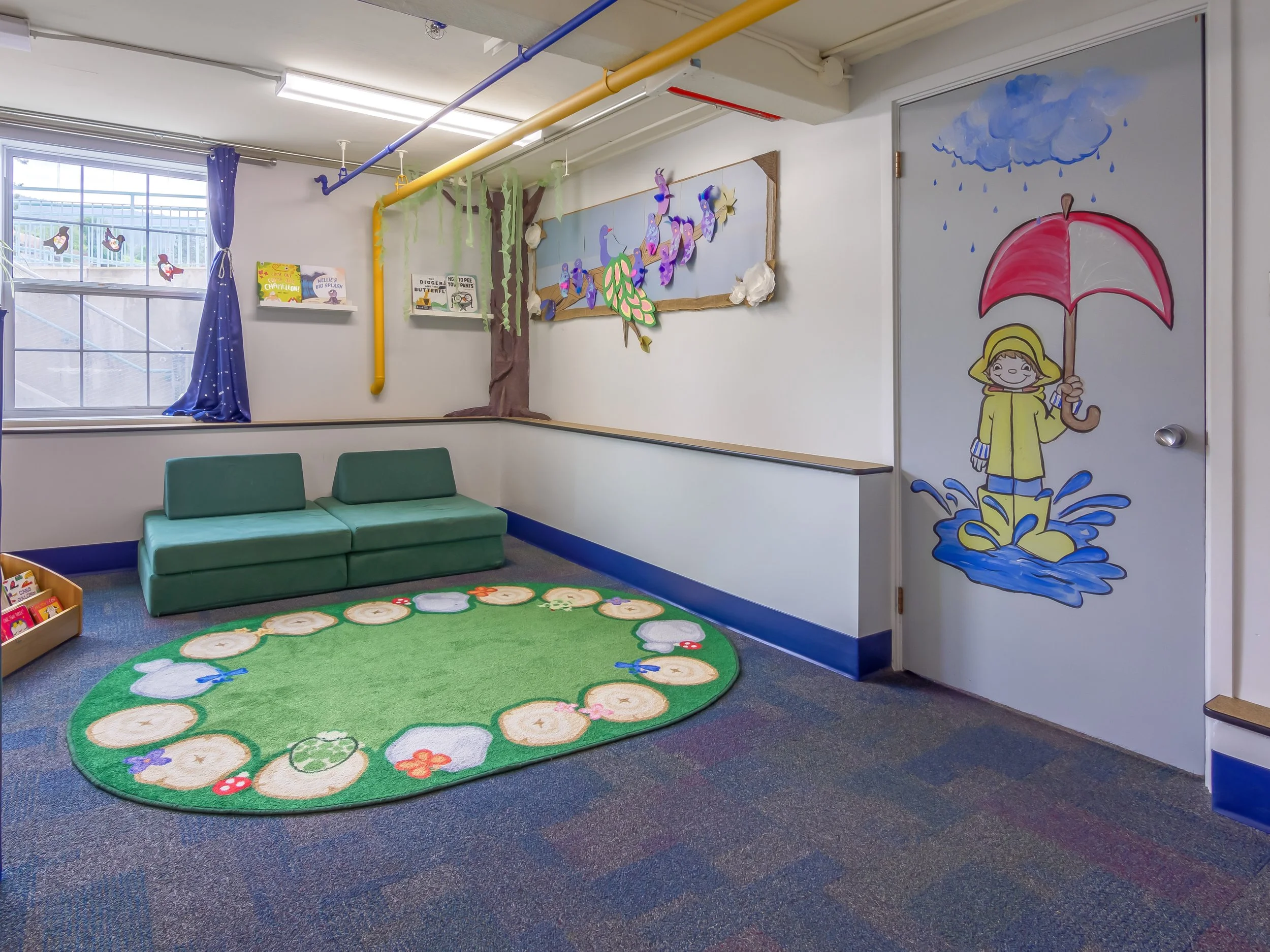 A colourful children's reading playroom with a green carpet shaped like a circle with animals and flowers, a green couch, stuffed animals, shelves with children's books, and painted walls with rain, flowers, butterflies, and a girl with an umbrella.