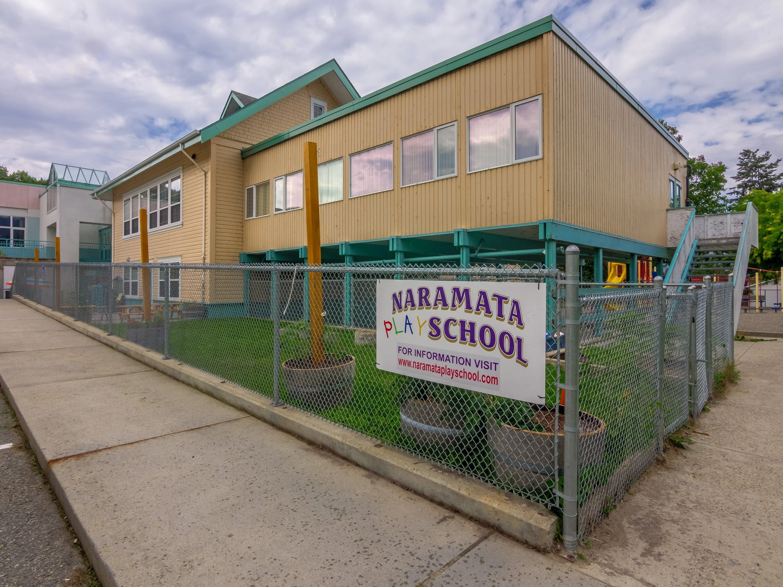 The Naramata Childcare Society building with a yellow exterior and teal roof, surrounded by a chain-link fence. There is a sign on the fence reading 'Naramata Play School' with a website link, and the yard has grass and large potted plants.