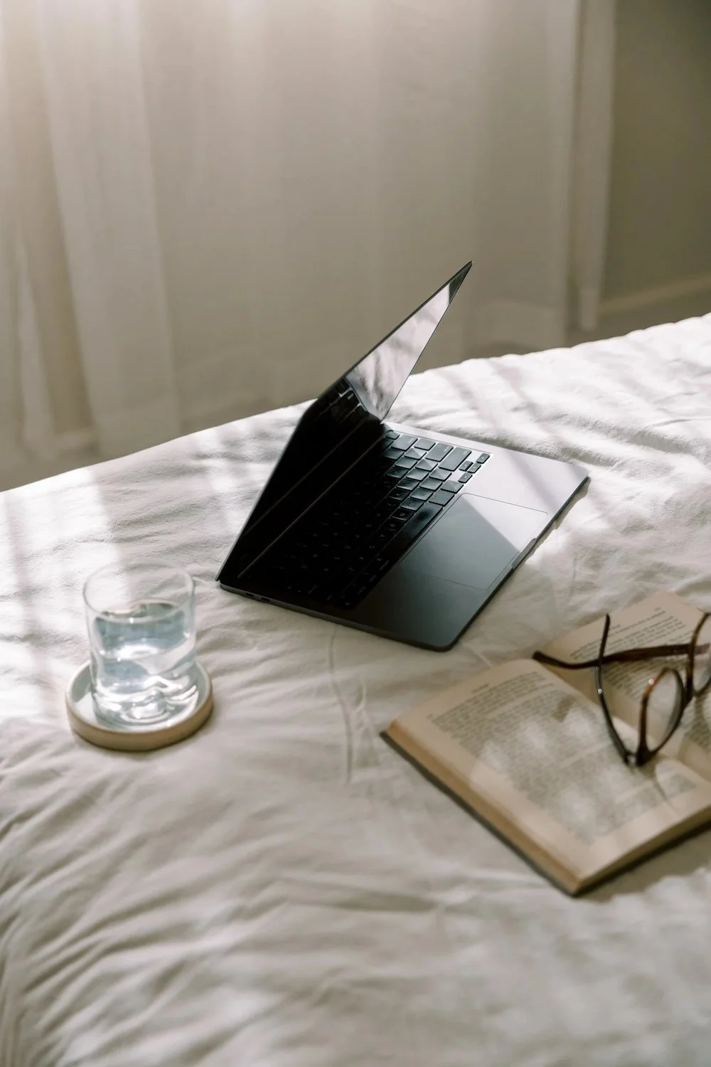 An open laptop sitting on a bed. There is a cup of water and an open book with reading glasses sitting nearby.