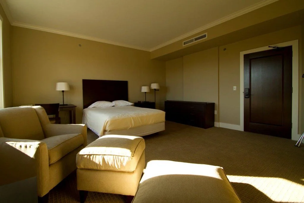 A hotel room with a bed, two nightstands with lamps, a reclining chair with an ottoman, and a door. Sunlight is streaming in through a window, creating shadows on the carpet.