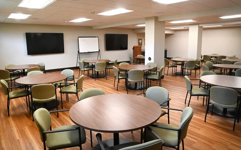 Conference room with round tables, chairs, two large screens, a whiteboard, and a podium.