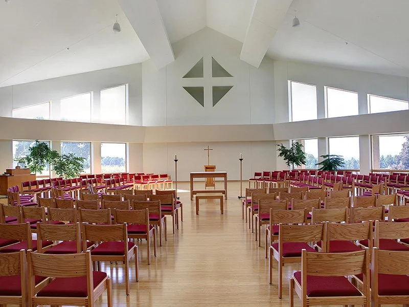 Interior of a church with wooden pews with red cushions, a cross on the wall, and large windows flood the space with natural light.