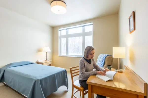 A woman sitting at a wooden desk in a bedroom, reading a book with a lamp, a bed with a blue bedspread, and a window in the background.