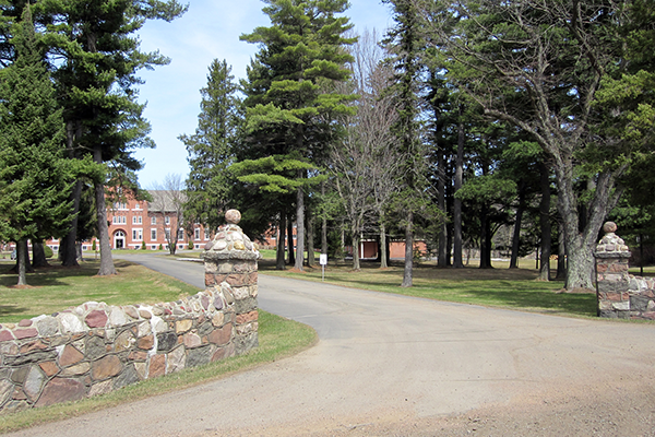 A gravel driveway with stone pillars on either side, leading to a historic brick building surrounded by tall trees and a well-maintained lawn.