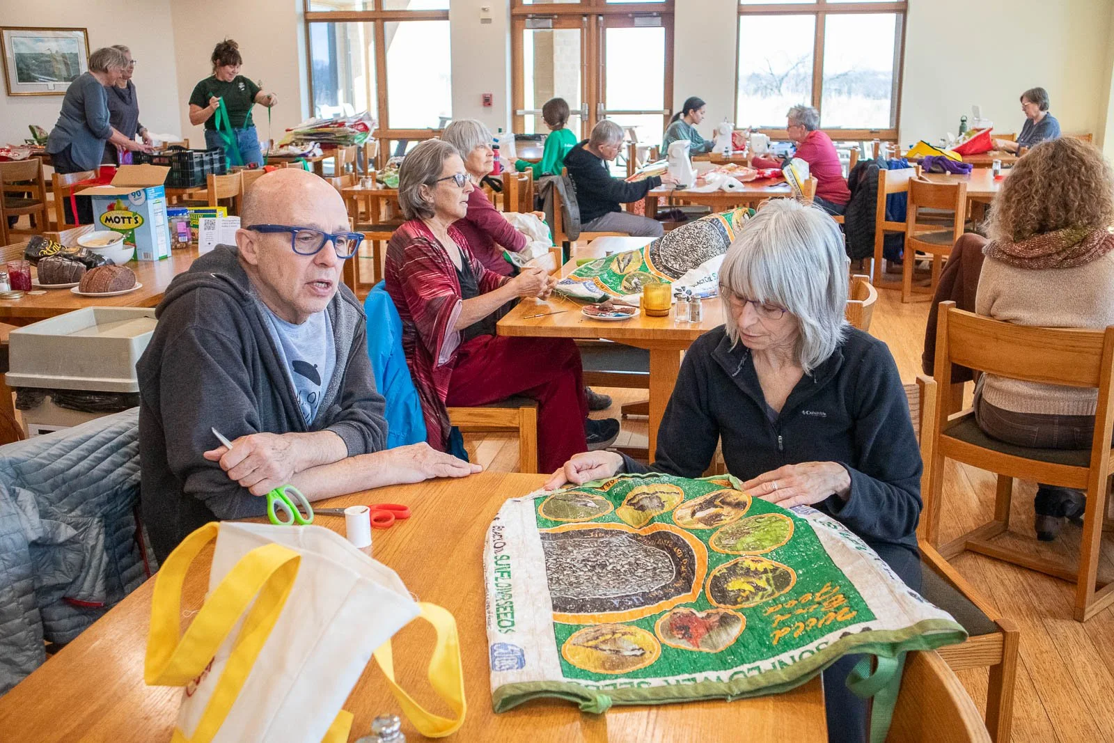 A group of people sitting at wooden tables in a room, engaged in quilting and crafting activities.