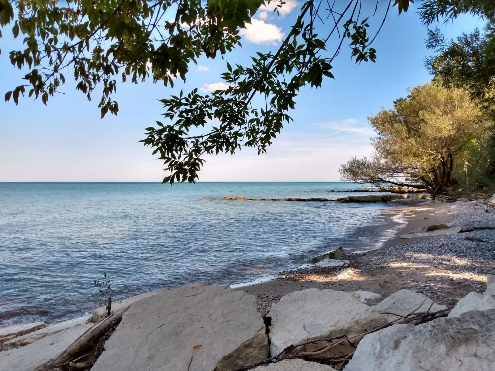 A peaceful beach scene with calm water, sandy and pebbly shore, green trees, and a blue sky with scattered clouds.