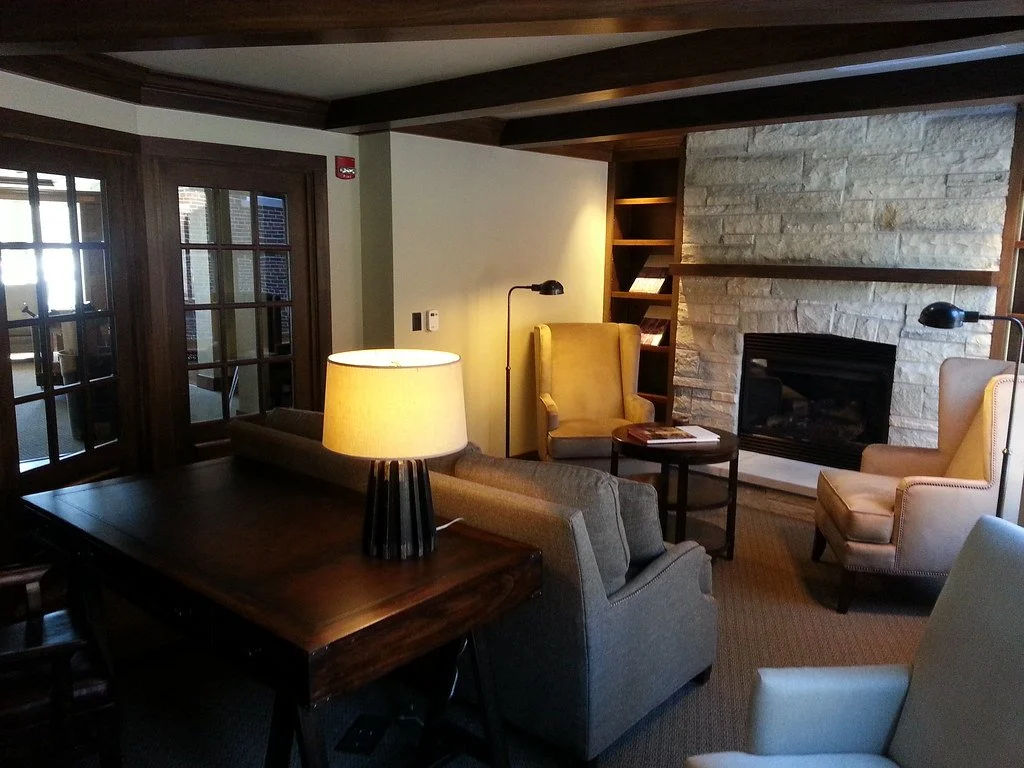 Cozy living room with beige and gray armchairs, a fireplace with a wooden mantel, a side table, a table lamp, and sliding glass doors leading outside.