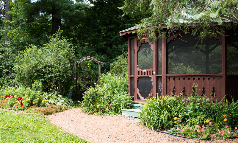 A garden next to a wooden screened gazebo with a garden path leading to a garden arch in the background.