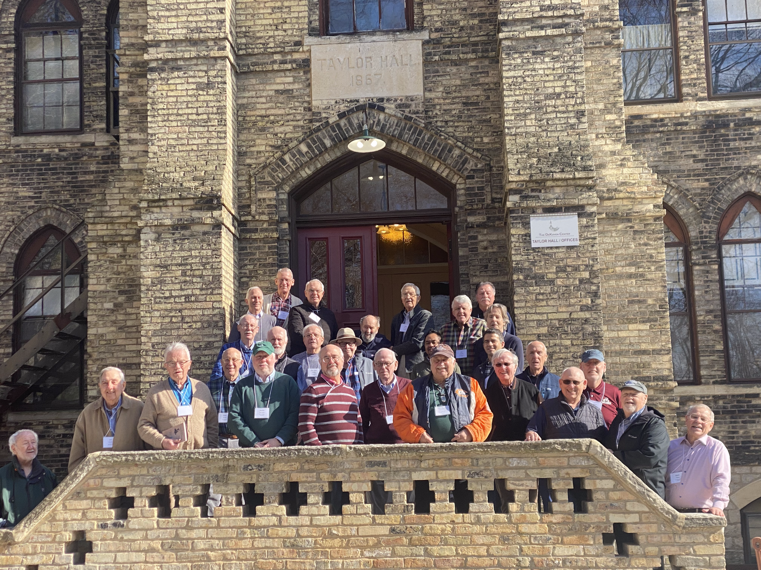 A group of elderly men and women standing on the steps of a historic brick building labeled Taylor Hall, 1867, posing for a group photo during a daytime gathering.