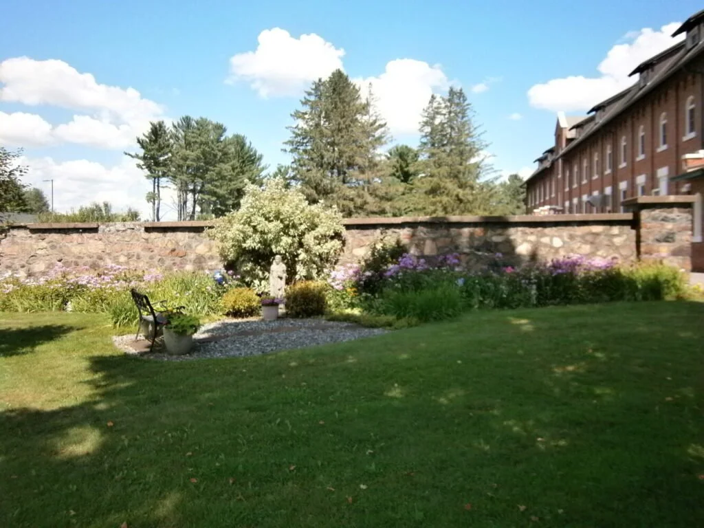 A landscaped yard with a stone wall, flower bed, a small tree, garden statues, benches, potted plants, and a well-maintained lawn, with trees and a building in the background under a partly cloudy sky.