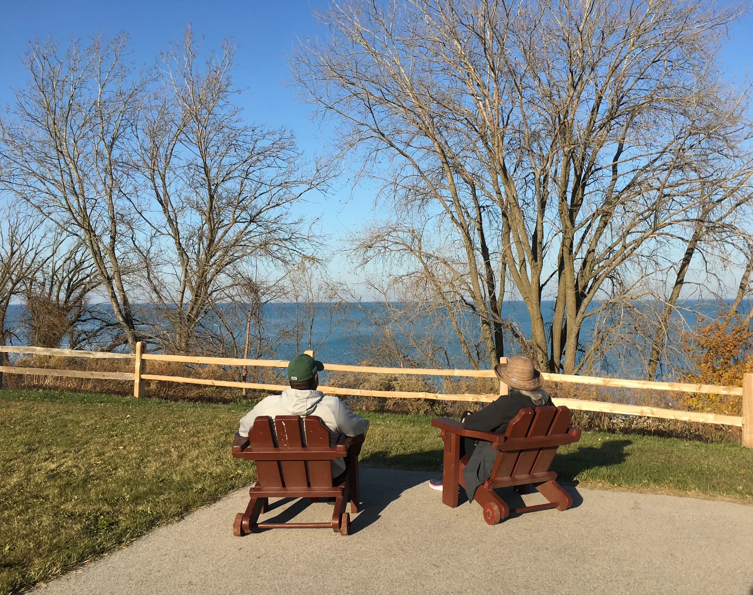 Two people sitting on wooden chairs facing a large tree by a fence, with a view of a body of water and a clear blue sky in the background.