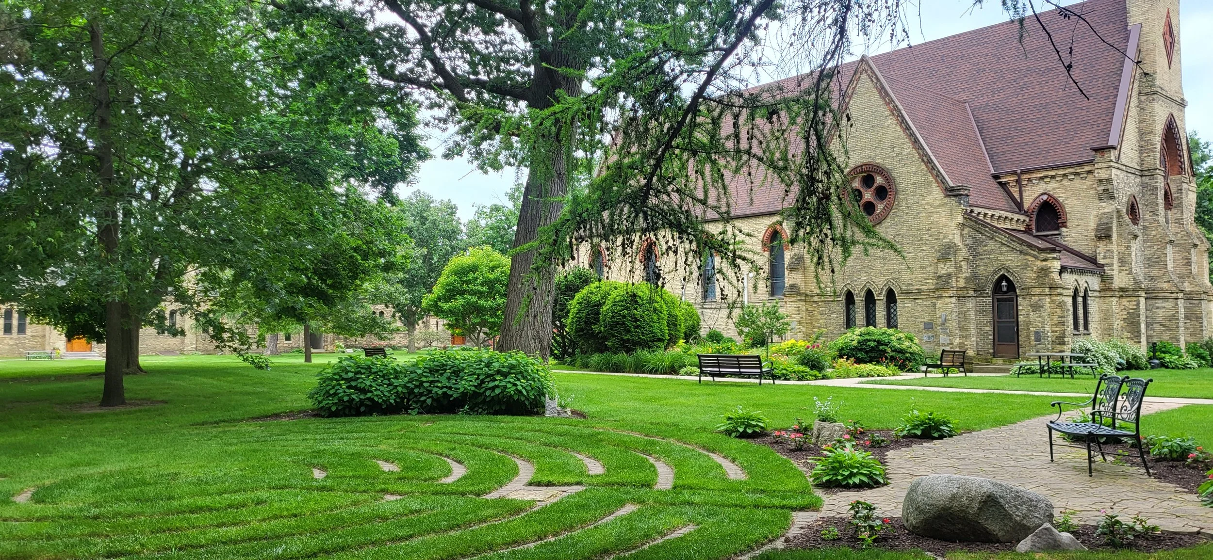 A historic stone church surrounded by a lush, well-maintained garden with benches, trees, and flower beds, under a cloudy sky.