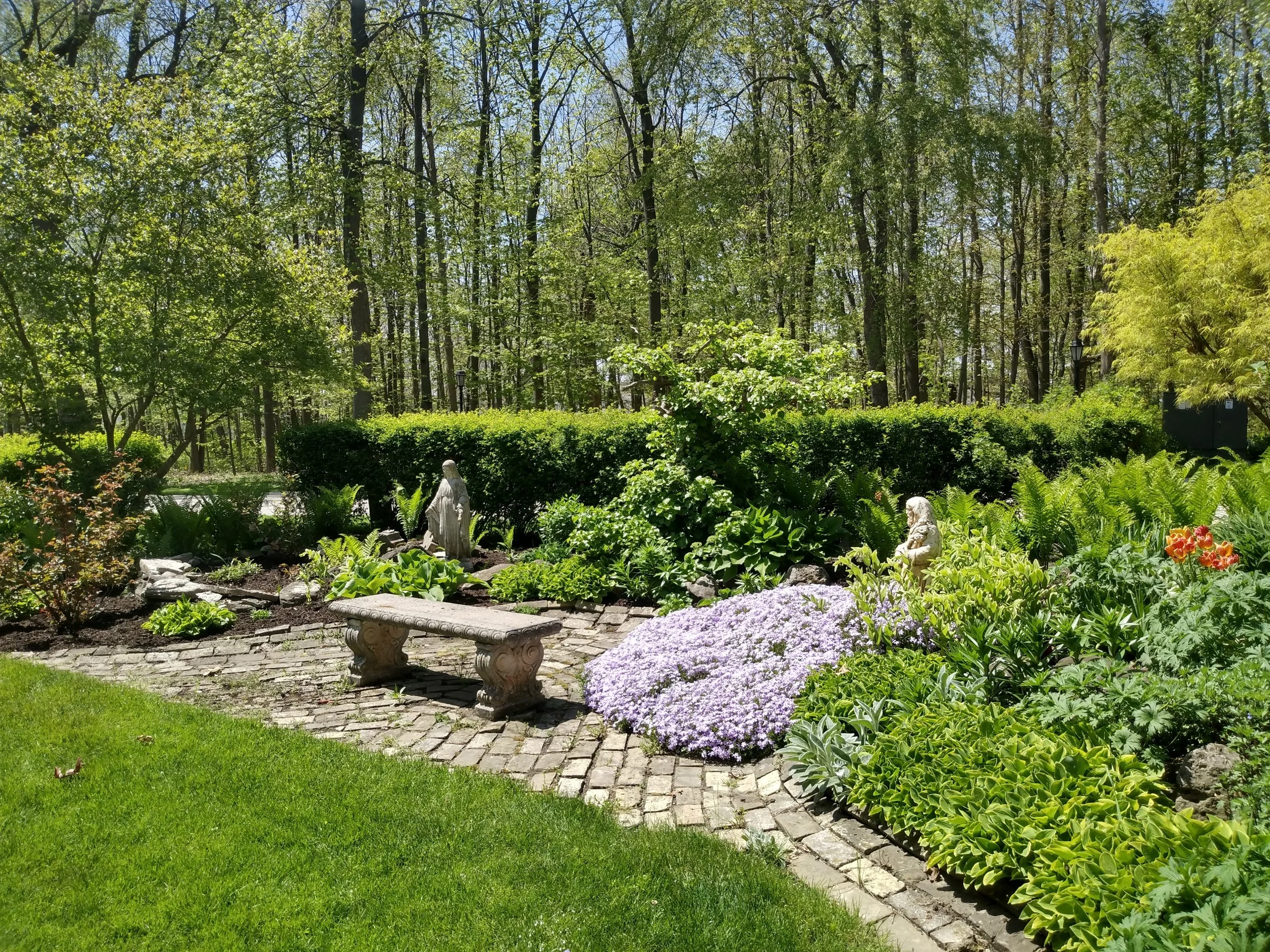 A lush garden with a paved brick path, a stone bench, statues, vibrant flowers, and dense greenery with trees in the background.