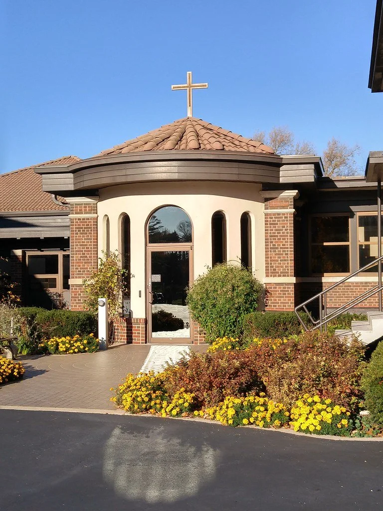 Church building with a cross on top, brick and stucco exterior, decorative bushes and flower beds at entrance, and clear blue sky.