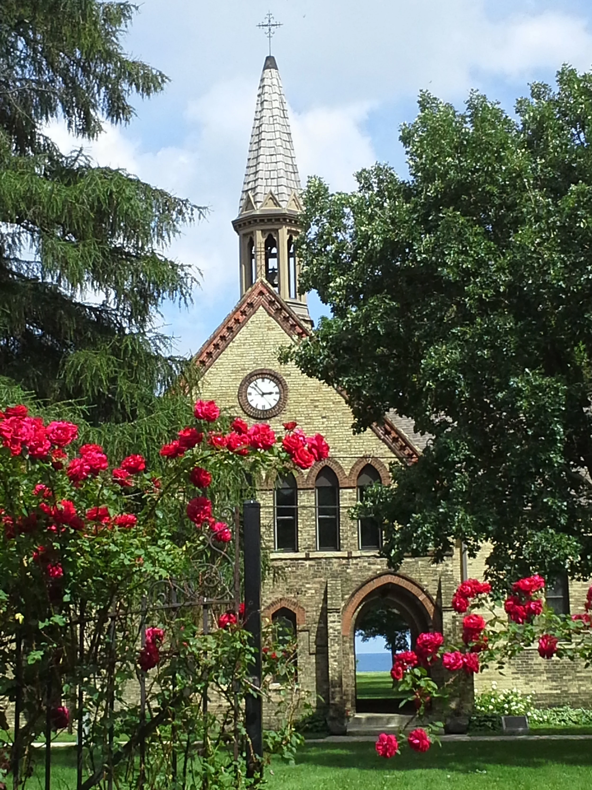 A Gothic stone building with a clock on its front, a tall steeple, and an arched entrance, surrounded by green trees and pink roses, with a cloudy sky in the background.