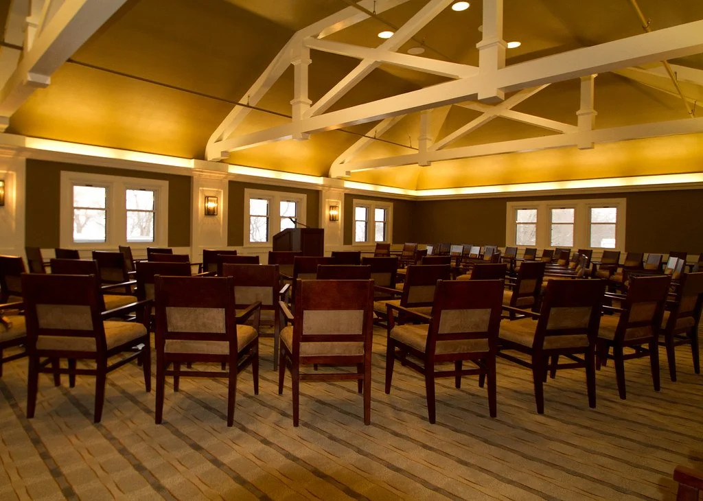 Empty conference room with wooden chairs arranged in circular seating, windows on the walls, and warm lighting.