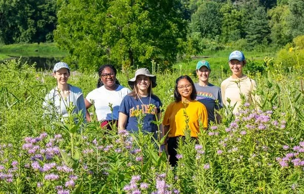 Six women standing in a field with purple flowers, tall green plants, trees, and a lake in the background.