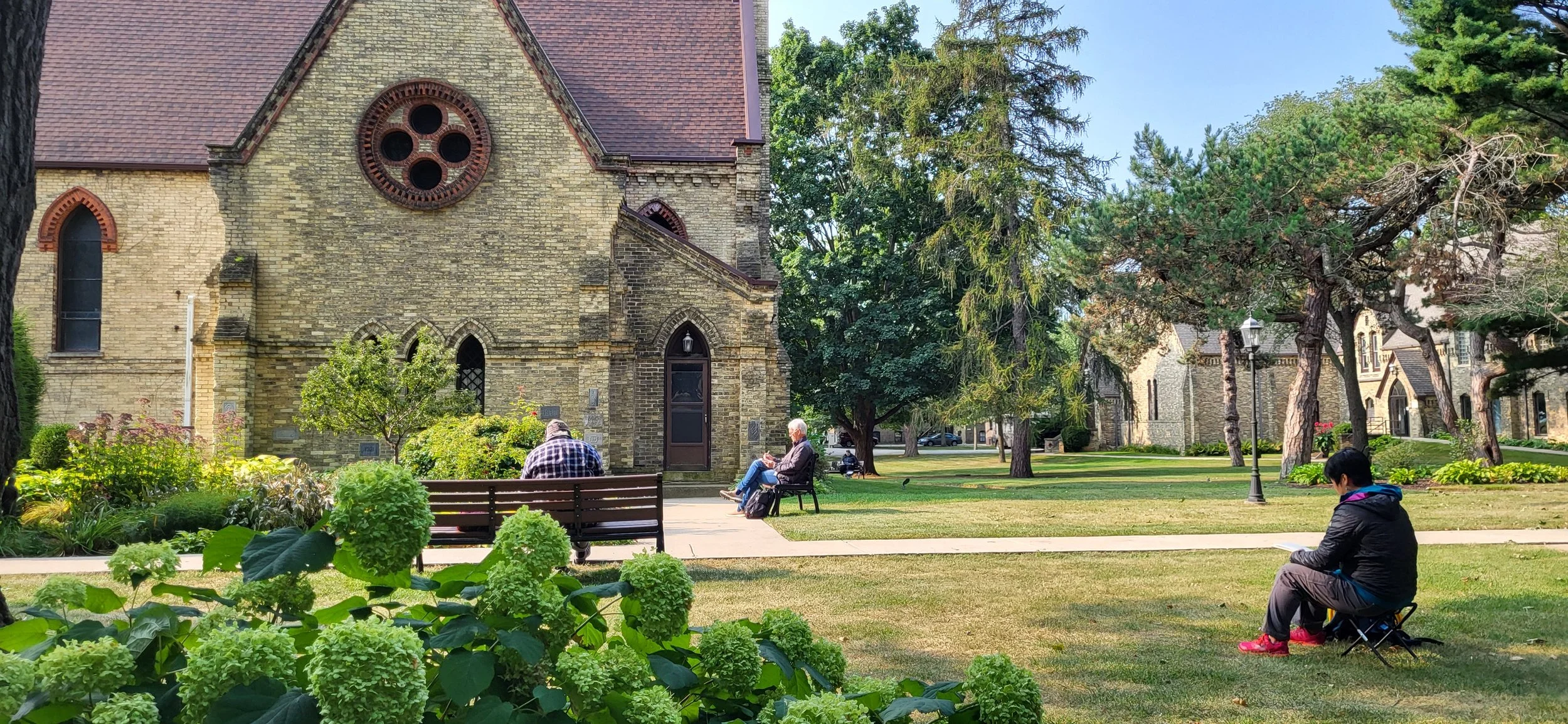 A peaceful park scene with an old brick church, green trees, and people sitting on benches and a small foldable chair, some reading or relaxing.