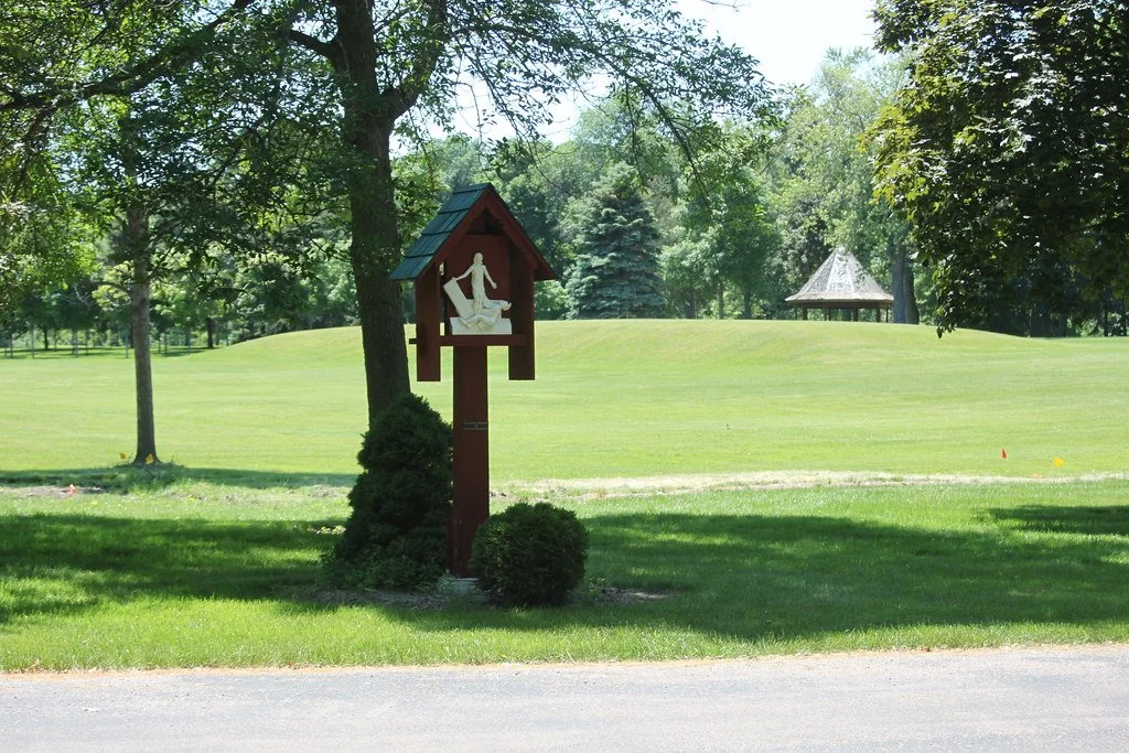 A peaceful park scene with a wooden religious shrine featuring a statue of Jesus, surrounded by manicured bushes and tall trees, with a grassy hill and a pavilion in the background.