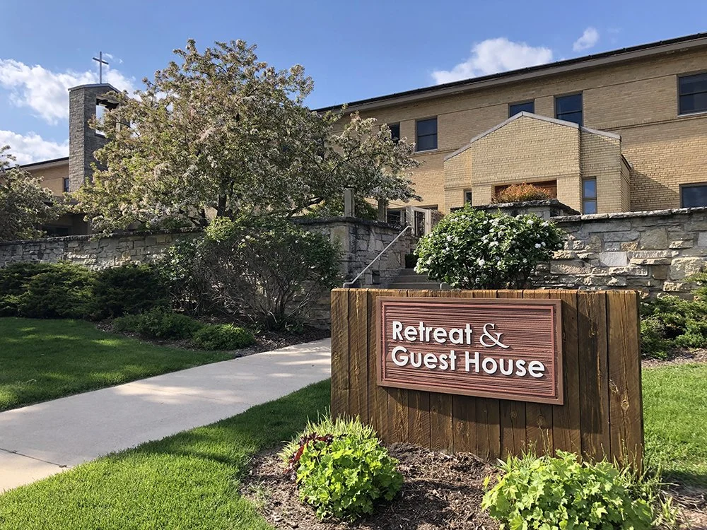 A sign reading 'Retreat & Guest House' in front of a large stone building with a brick exterior, surrounded by well-maintained lawn, shrubs, and flowering trees under a blue sky.