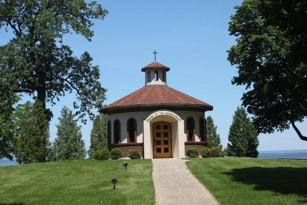 Small chapel with a red conical roof and a cross on top, surrounded by green trees and a neatly maintained lawn, located near a body of water.