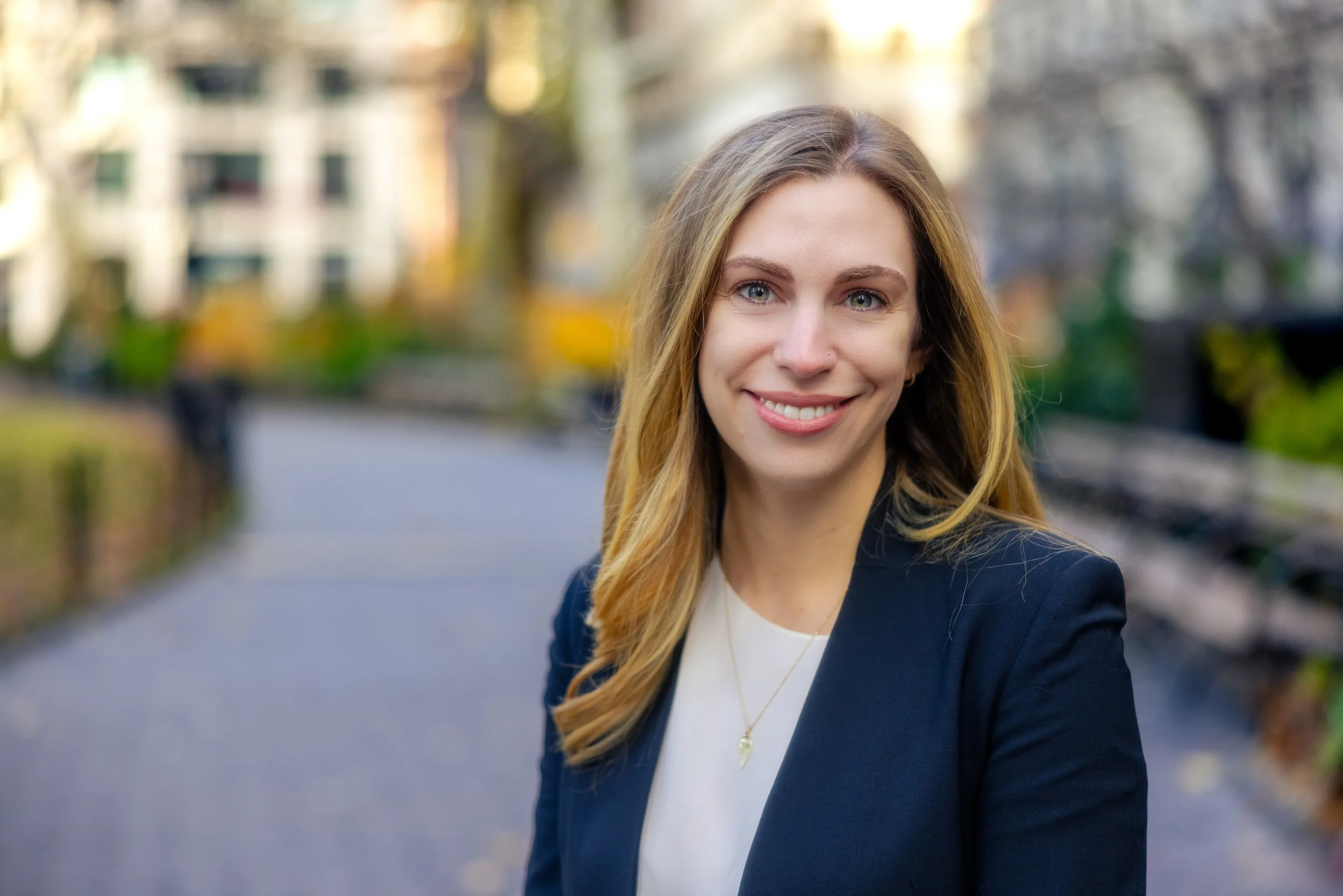 A smiling woman with long blonde hair, wearing a dark blazer and a white top, standing outdoors in a park with blurred trees and buildings in the background.