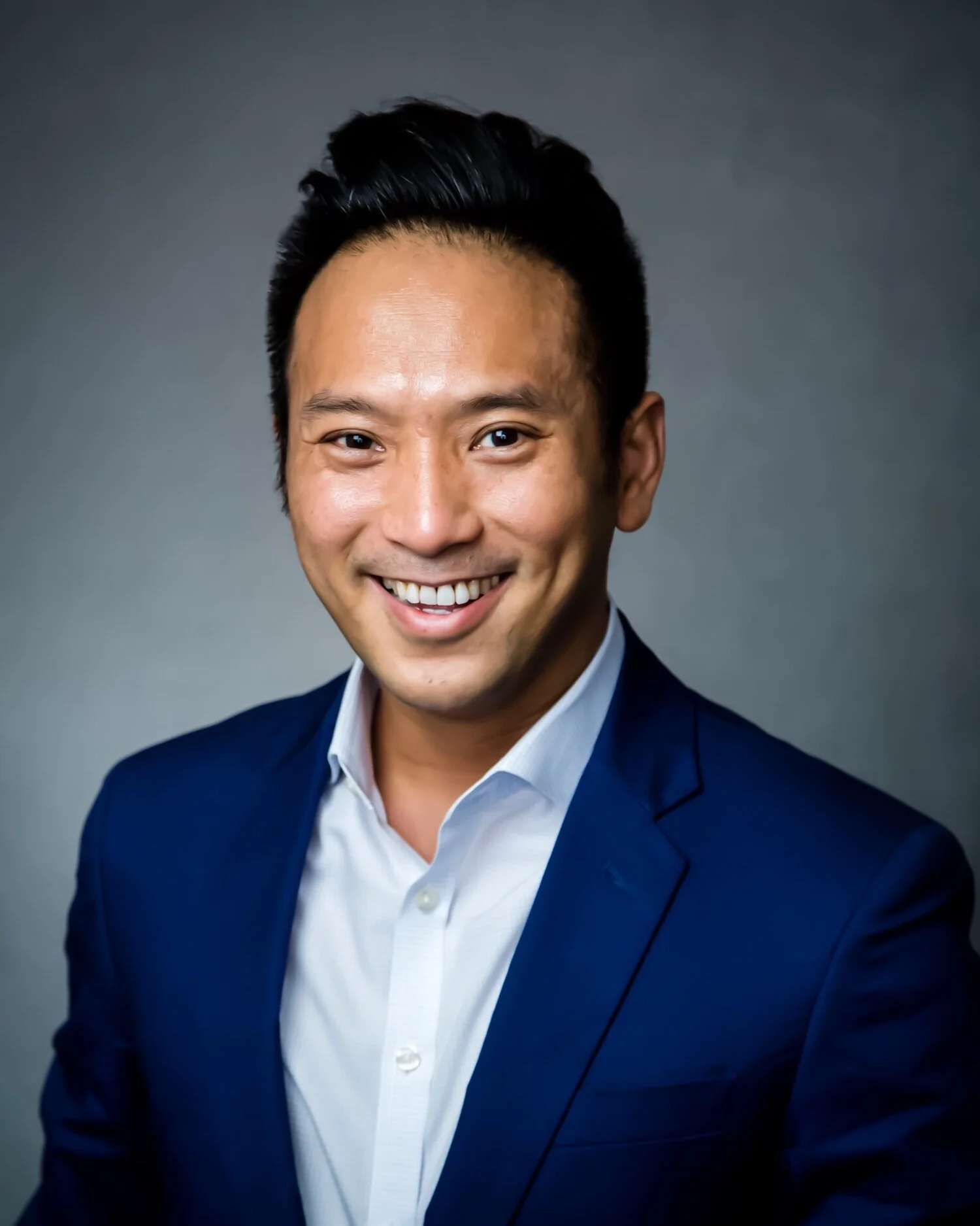 Professional headshot of a smiling man with short black hair, wearing a white dress shirt and blue blazer, against a gray background.