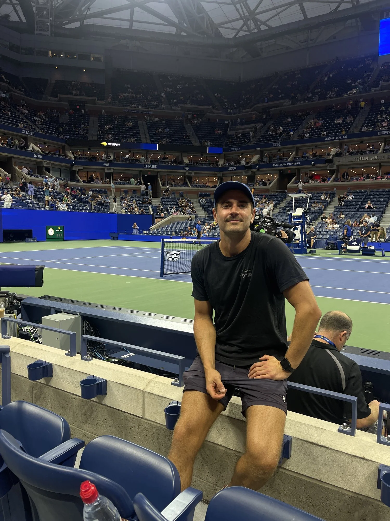 A man sitting on a bench at a tennis stadium, wearing a black t-shirt, shorts, and a blue cap, with tennis courts and spectators in the background.