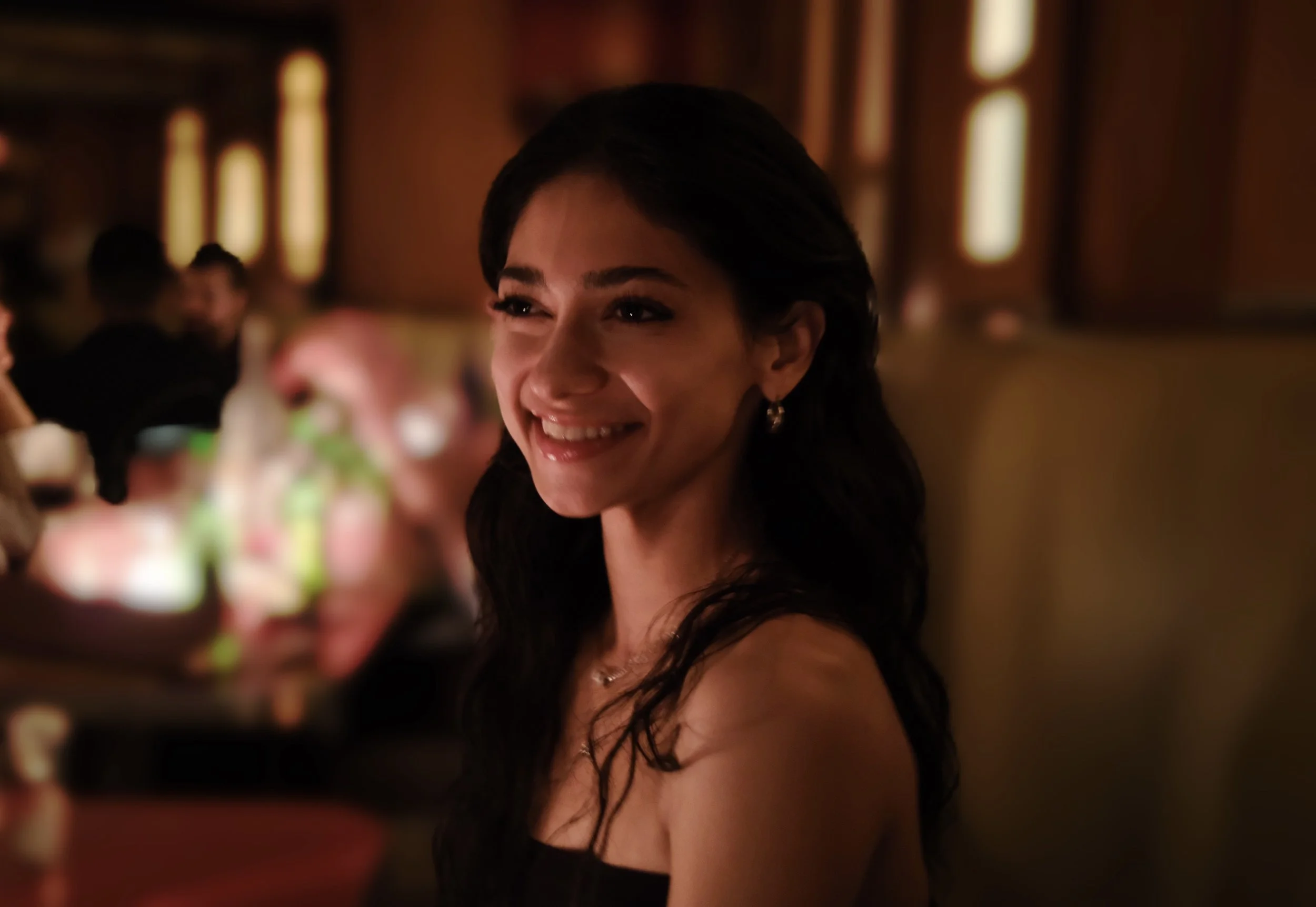 A smiling woman with dark hair and earrings in a dimly lit indoor setting with other people in the background.