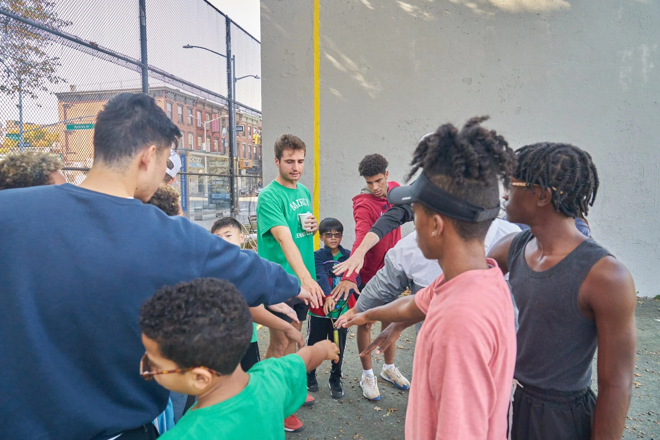 A group of diverse children and teenagers gathered in a circle on a basketball court, participating in a team-building or coaching activity, with one adult coach in a green shirt.
