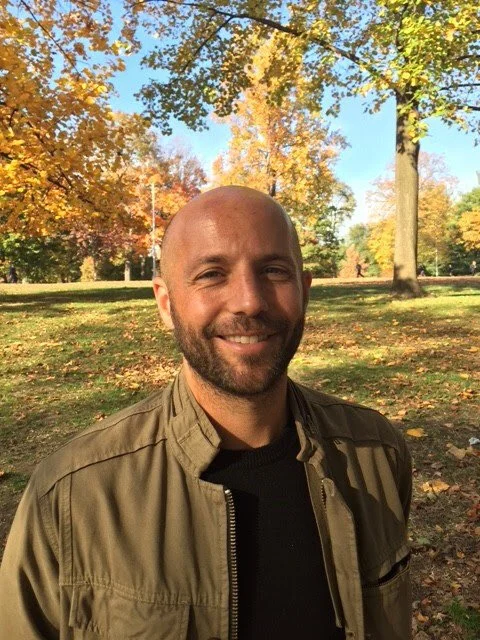 A smiling man with a beard and a shaved head standing outdoors in a park during autumn, with trees showing fall foliage behind him.