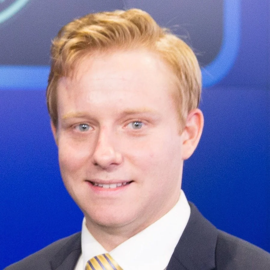 A young man with light skin, ginger hair, blue eyes, wearing a dark suit, white shirt, and a yellow striped tie, in front of a blue background with a partial logo.
