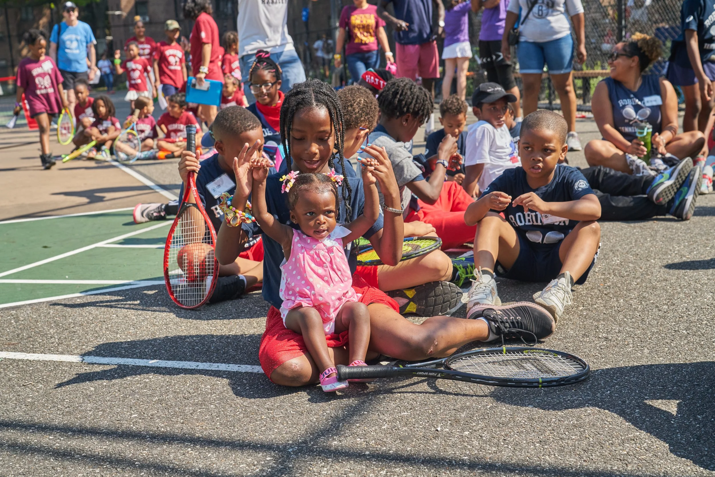 Group of children and adults sitting and lying on the ground at an outdoor tennis court during daytime, some holding tennis racquets, with more children standing in the background.