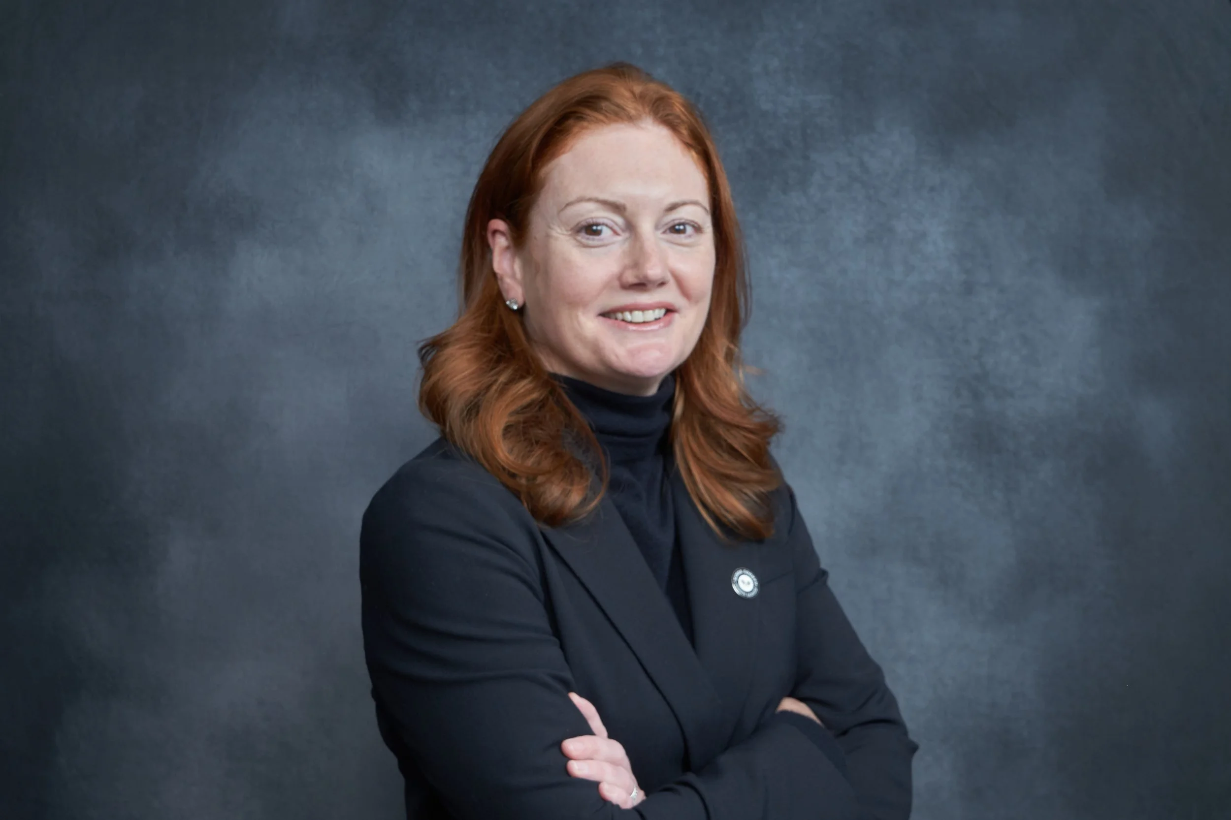 A woman with red hair, wearing a black blazer and turtleneck, smiling with arms crossed against a dark grey background.