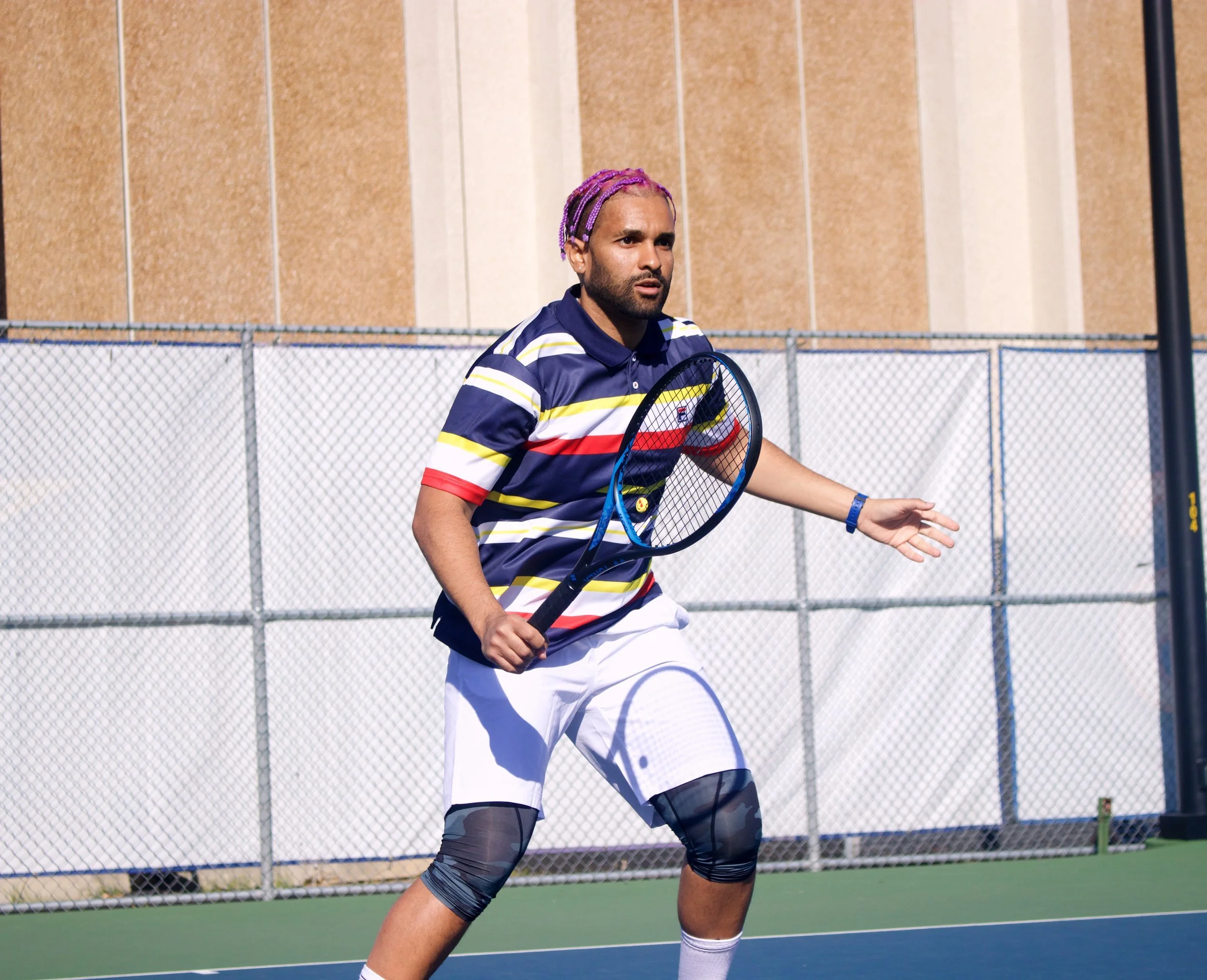 A man with purple braided hair playing tennis on an outdoor court, wearing a striped polo shirt, white shorts, and knee-length compression sleeves.