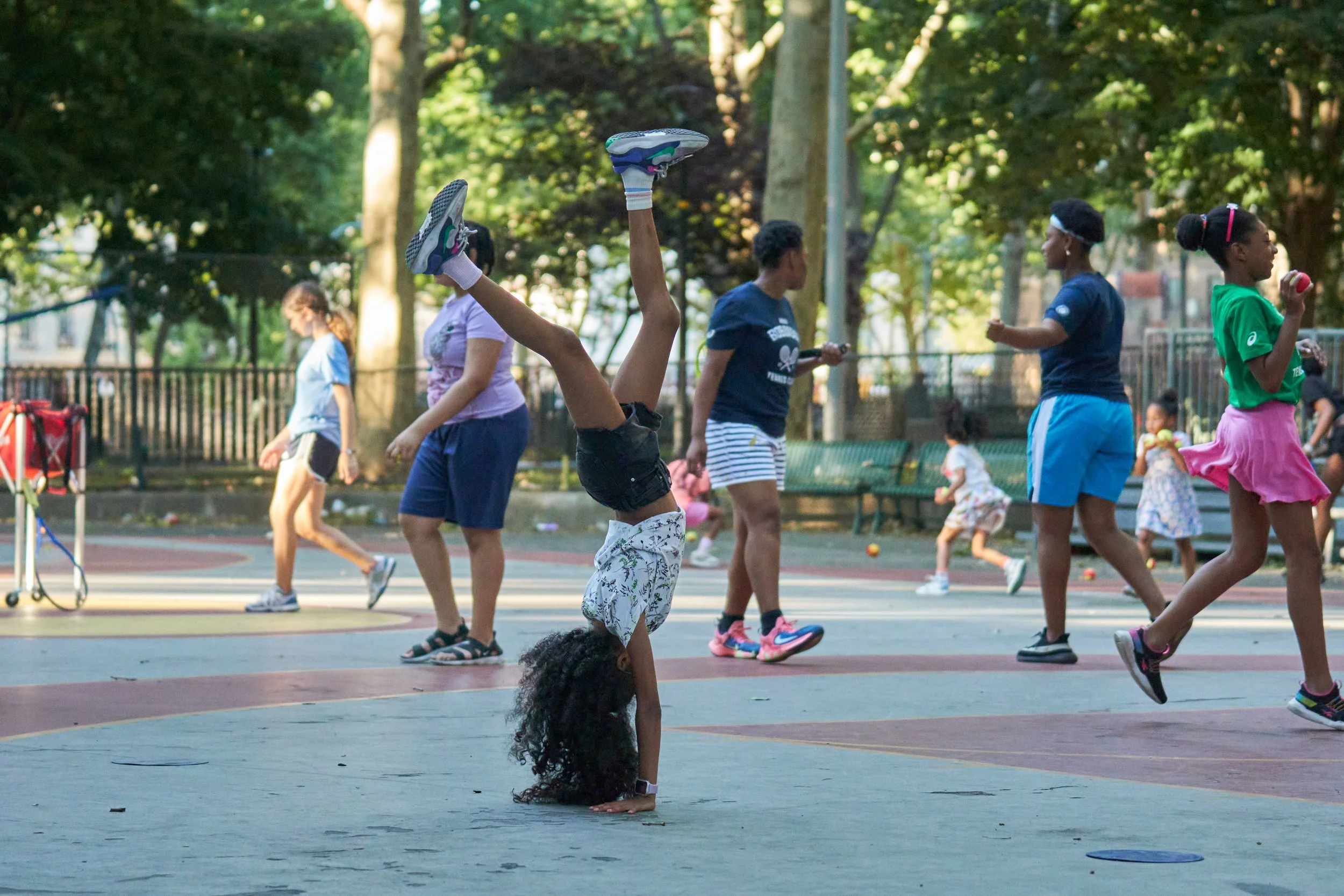 Children playing at a park, including one performing a handstand and others running or walking, with trees and benches in the background.