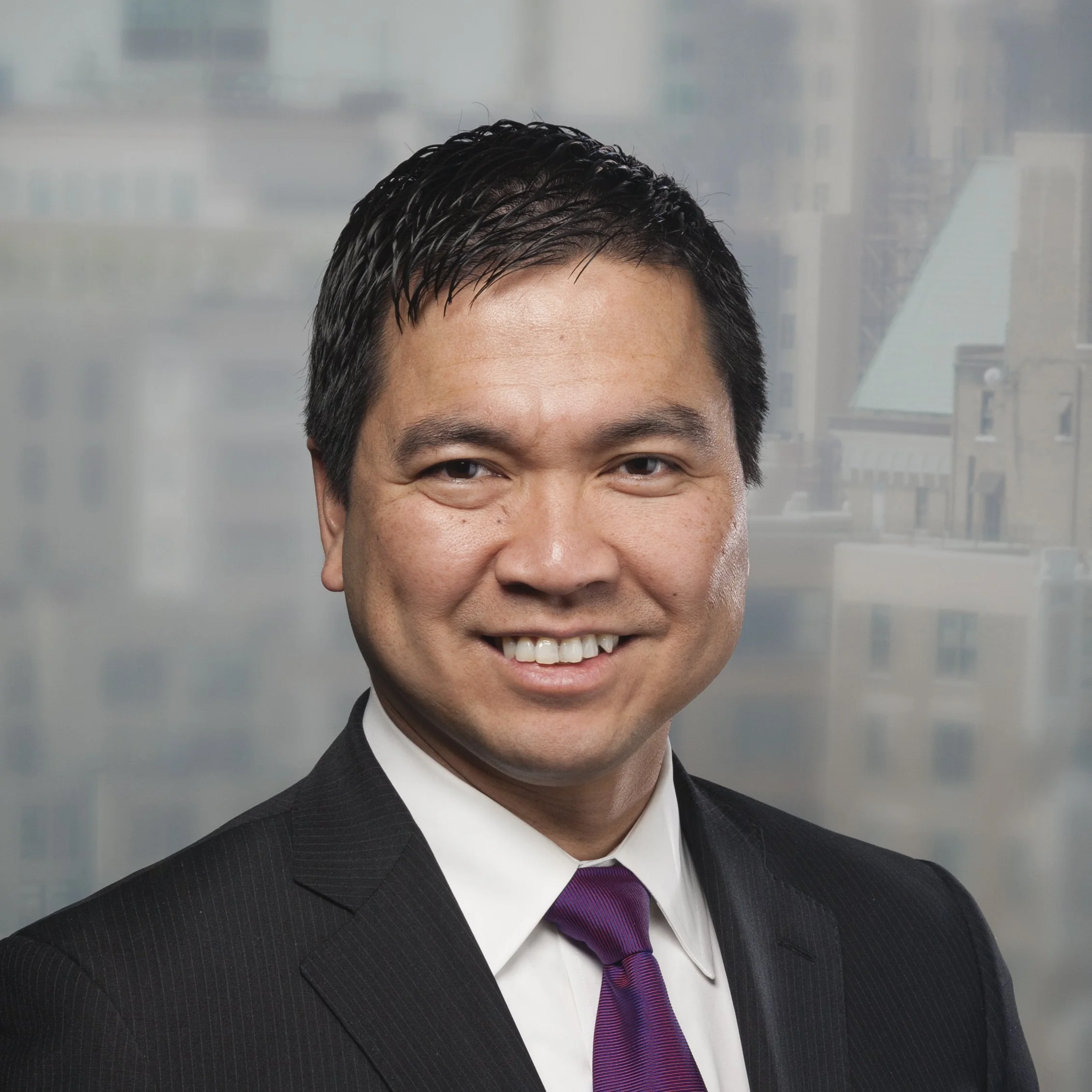 Professional headshot of a smiling man with dark hair, wearing a suit and purple tie, in front of a cityscape with buildings and a cloudy sky.