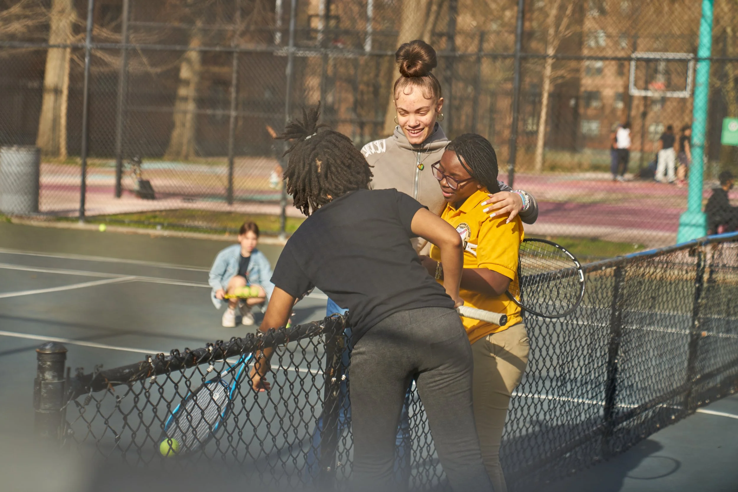 Three girls embracing on a tennis court, smiling, with a young girl sitting in the background holding tennis balls.