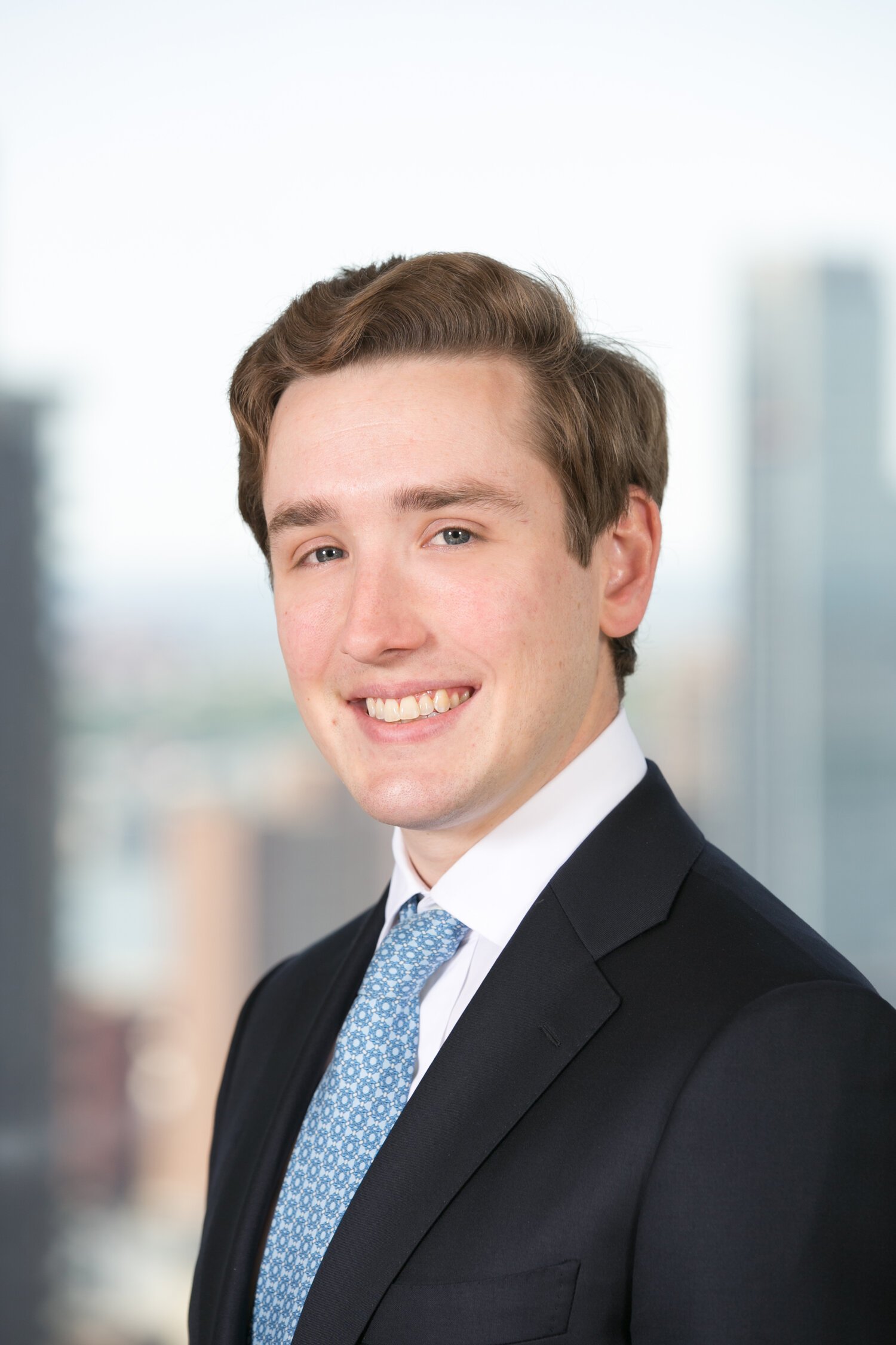 Young man with brown hair in a suit and tie, smiling in a professional headshot with a blurred cityscape background.